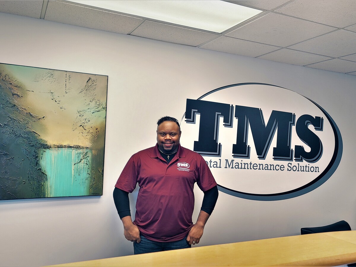 a man smiling while he stands with the TMS logo on the wall behind him